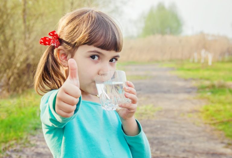 girl outside drinking a glass of water and showing the thumbs up sign