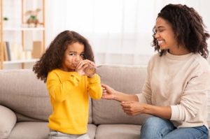 girl drinking water with mom smiling at her while both sit on couch